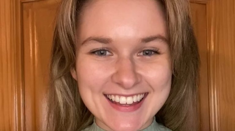 A woman with long, light brown hair smiles at the camera, standing in front of a wooden door whilst wearing a light green polo-neck top.
