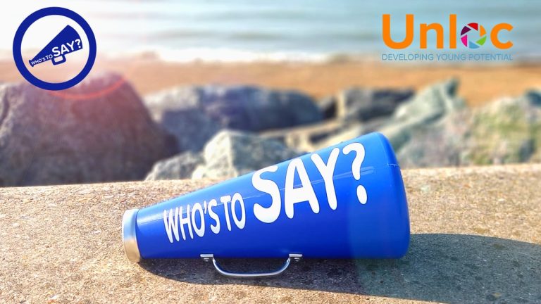 A blue megaphone labelled WHO'S TO SAY? rests on rocks by a beach, with the Unloc logo and slogan DEVELOPING YOUNG POTENTIAL in the upper right corner.