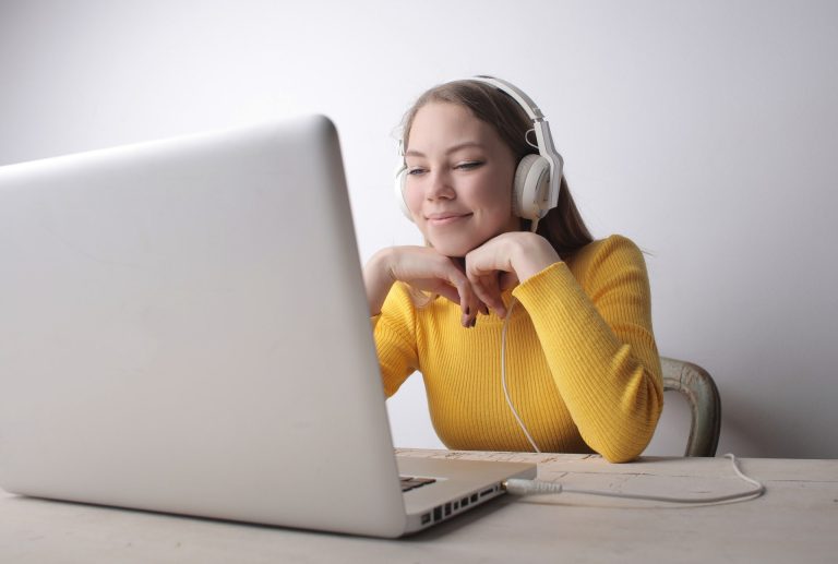 A person wearing a yellow jumper and headphones sits at a table, looking at a laptop screen with a slight smile.